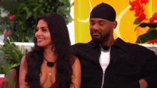 a man and a woman are sitting next to each other and the woman is wearing a necklace with the letter s on it