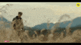 a man in a military uniform is standing in a field of tall grass with mountains in the background .