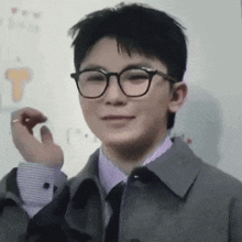 a young man wearing glasses and a jacket is standing in front of a whiteboard .