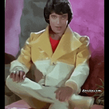a man in a yellow jacket sits on a couch with anshu.com in the corner