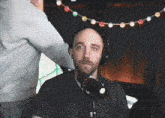 a man wearing headphones sitting in front of a microphone with a string of lights hanging from the ceiling behind him