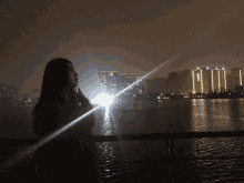 a woman stands in front of a body of water with buildings in the background and a light shining on her face
