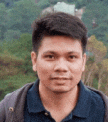 a man wearing a blue shirt and a brown jacket is standing in front of a forest .