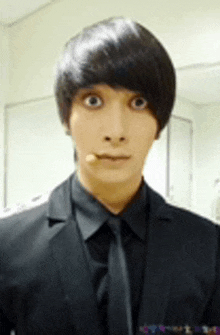 a young man wearing a black suit and tie looks at the camera