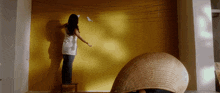 a woman is standing on a small stool painting a yellow wall