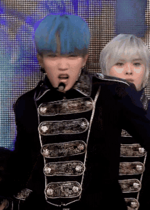 a man with blue hair is singing into a microphone while another man looks on