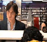 a man in a suit sits at a counter in front of a sign that says " new "