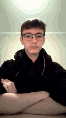 a young man wearing glasses and a black hoodie looks at the camera with his arms crossed