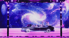 a group of people sitting in a car on a stage with a galaxy in the background