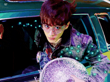 a man wearing glasses and a denim jacket is holding a silver object in front of a car