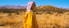 a woman in a yellow coat stands in a desert with mountains in the background