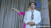 a man in a white suit and tie is standing next to a woman in a pink shirt and pointing .