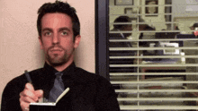 a man in a black shirt and tie is holding a notebook and pen .