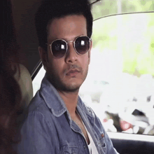 a man wearing sunglasses and a denim jacket looks at the camera