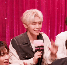 a man is holding a microphone in front of a pink curtain while sitting in front of a group of people .