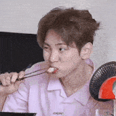 a young man in a pink shirt is eating food with chopsticks and a fan in the background