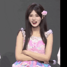 a woman in a pink dress with a flower in her hair is smiling with her arms crossed .