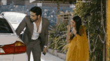 a man in a suit and a woman in a yellow sari are standing next to a white car .