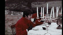 a man in a red coat is drinking from a cup at a table with candles