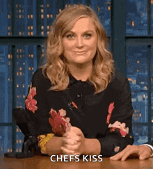 a woman in a floral shirt is sitting at a table with her hands folded and says chefs kiss .
