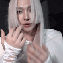 a woman with long white hair is wearing white gloves and making a heart shape with her hands .