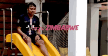 a man sits on a yellow slide with zimbabwe written on the wall