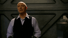 a bald man in a white shirt and black vest stands in front of a wall