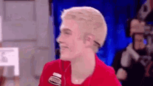 a young man with blonde hair is smiling and wearing a red jacket .