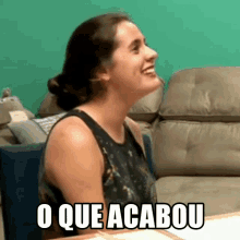 a woman is sitting on a couch and smiling with the words o que acabou written on the screen behind her .