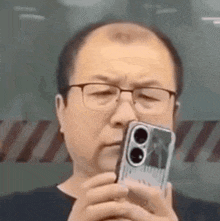 a bald man wearing glasses is taking a picture of himself with his cell phone .