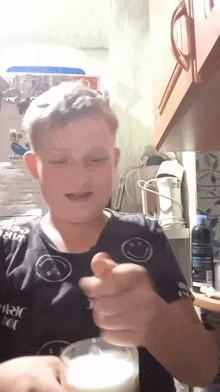 a boy wearing a black shirt with a smiley face on it holds a glass of milk