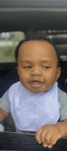 a baby wearing a bib and a gray shirt looks out of a car window