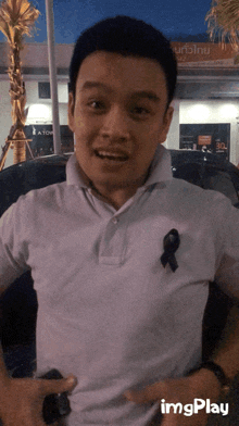 a man wearing a white shirt with a black ribbon on his chest is standing in front of a sign that says imgplay