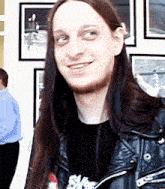 a man with long hair wearing a black shirt and a black leather jacket smiles