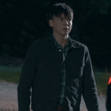 a young man wearing a black jacket and plaid shirt is walking down a dirt road at night