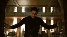 a man in a black jacket is standing in a room with his arms outstretched in front of a window .