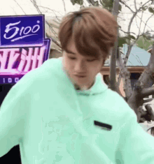 a young man wearing a green hoodie is standing in front of a tree .