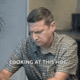 a man in a plaid shirt is sitting at a desk with the words looking at this hog above him