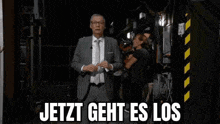 a man in a suit and tie is standing in a dark room with the words `` jetzt geht es los '' behind him .