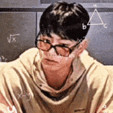 a young man wearing glasses is sitting in front of a chalkboard with mathematical equations written on it .