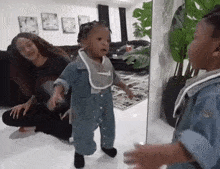 a baby is standing in front of a mirror in a living room while a woman sits on the floor .
