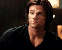 a man with long hair is wearing a black shirt and looking at the camera with a serious look on his face .