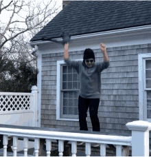 a man in a gray sweater and black hat stands on a white railing in front of a house