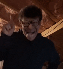 a man wearing glasses and a black sweater is making a funny face while standing in an attic .