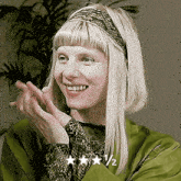 a woman with blonde hair wearing a headband and a green jacket is smiling and clapping her hands