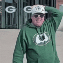 a man wearing a green hoodie , sunglasses and a hat is standing in front of a green bay packers sign .