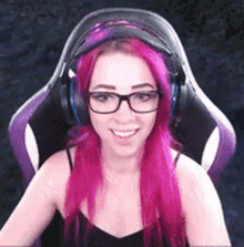 a woman with pink hair and glasses is sitting in a purple chair wearing headphones .