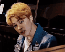 a young man with blonde hair is wearing a blue leather jacket and a white t-shirt .