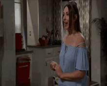 a woman wearing a blue off the shoulder top is standing in a kitchen