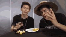 two young men are sitting at a table with a plate of food and one of them is wearing a straw hat .